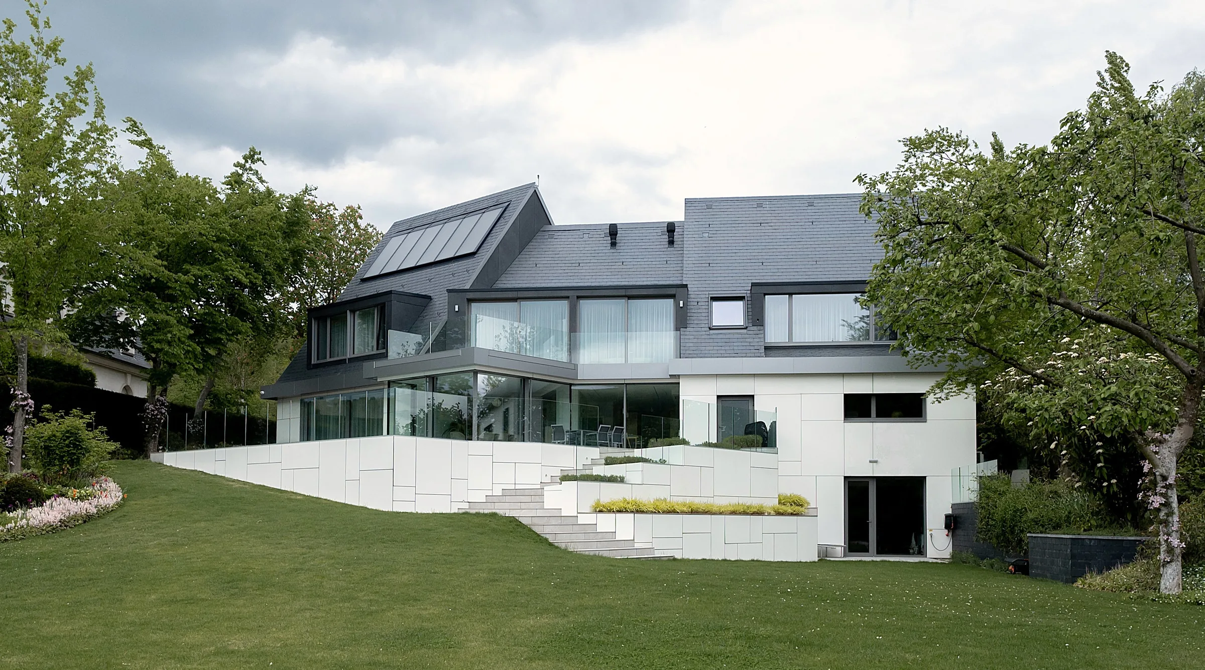 Extension de maison à Waldbredimus : volumes en panneaux blancs et ardoise, terrasses vitrées au-dessus d'un jardin paysager